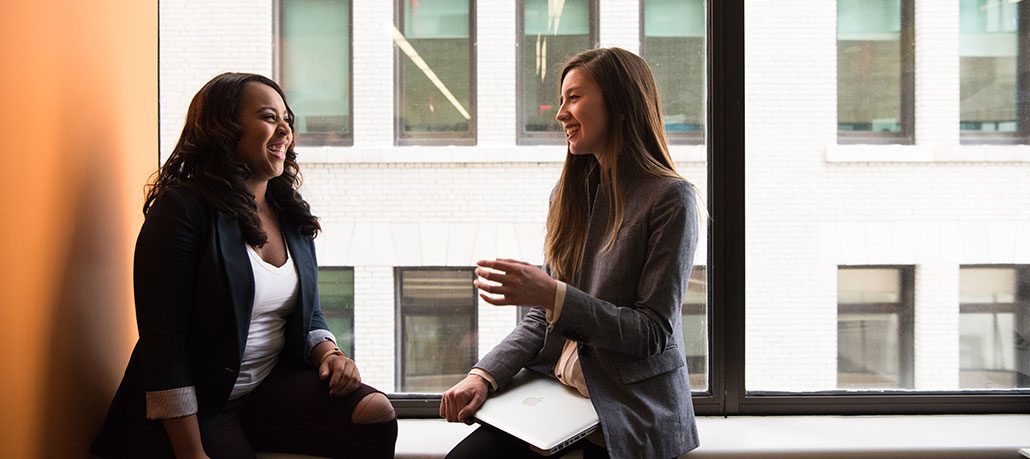 Two women in suits talking happily near a window in an office building