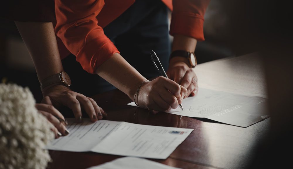 Close up of two people's arms as they lean over a desk to sign paperwork